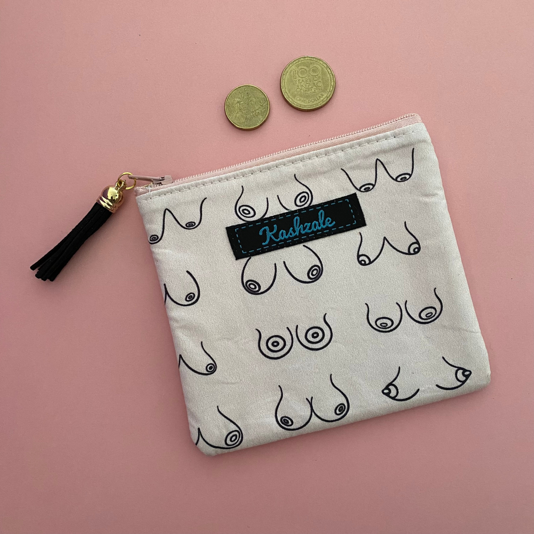 A small coin purse perfect for coins and small essentials featuring various sizes of breasts on light pink fabric and a tassel on the zipper.