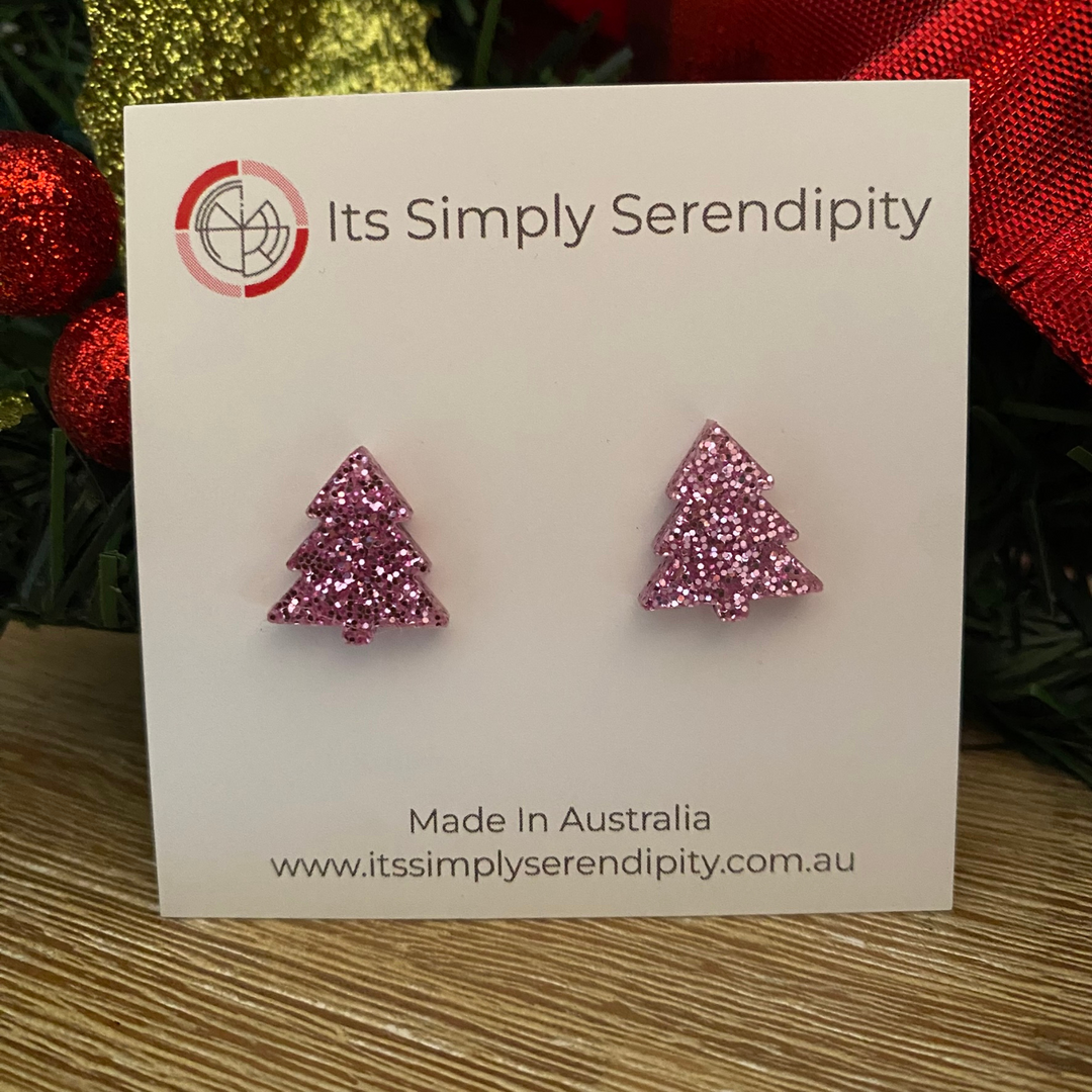 Pink glittery tree-shaped earrings on a 'Its Simply Serendipity' card with Christmas decorations in the background.