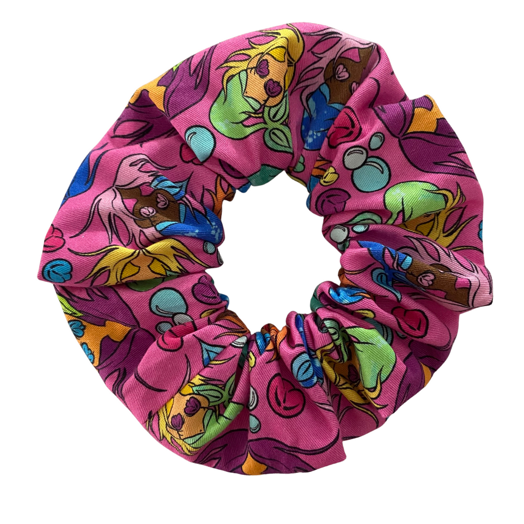 A scrunchie hair tie handmade with fabric by Kasey Rainbow with images of colourful mermaids on a pink background.