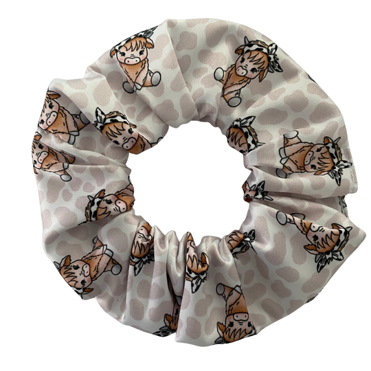A scrunchie with images of a baby cow wearing a bow headband on a light brown spotted fabric.