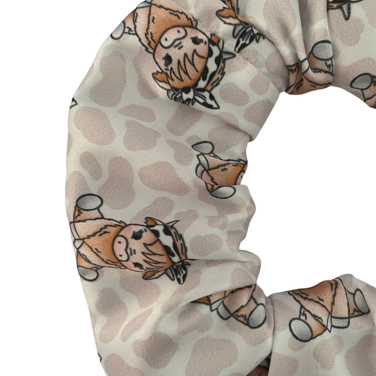 Close up view of a scrunchie with images of a baby cow wearing a bow headband on a light brown spotted fabric.