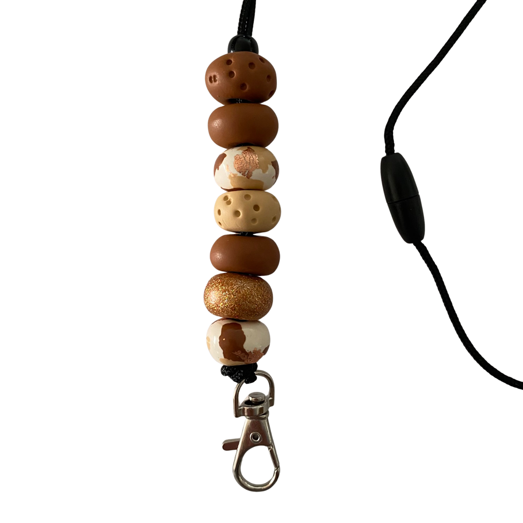 Handmade polymer clay lanyard with seven hand-rolled beads in brown, cream and intricate patterns accented with gold foil. Threaded on a nylon cord with a breakaway safety clasp and a silver-plated clip.
