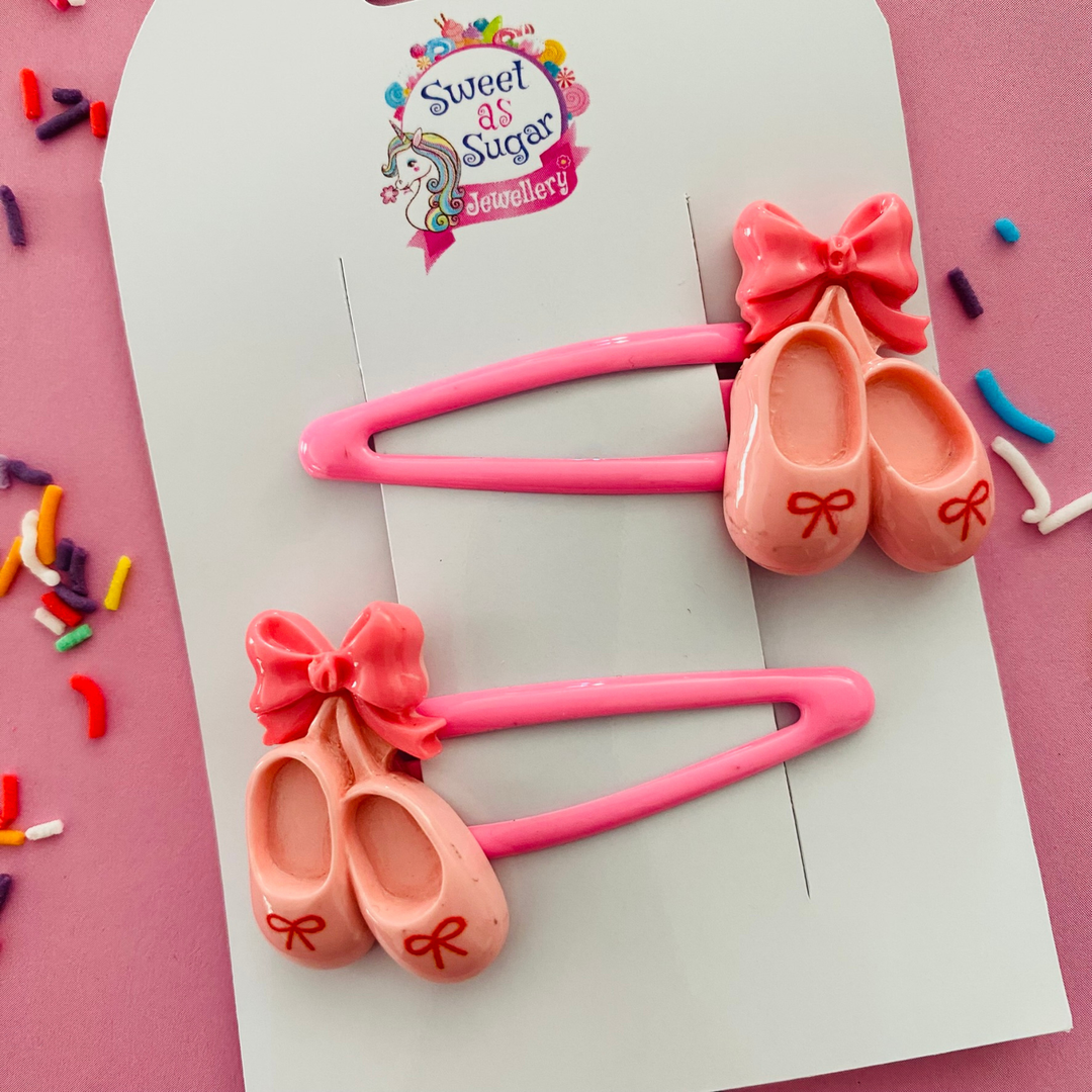 A pair of pink snap clips featuring pink ballet shoes adorned with a pink bow.