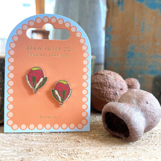 A pair of Banksia enamel stud earrings on a branded card with natural elements in the background.