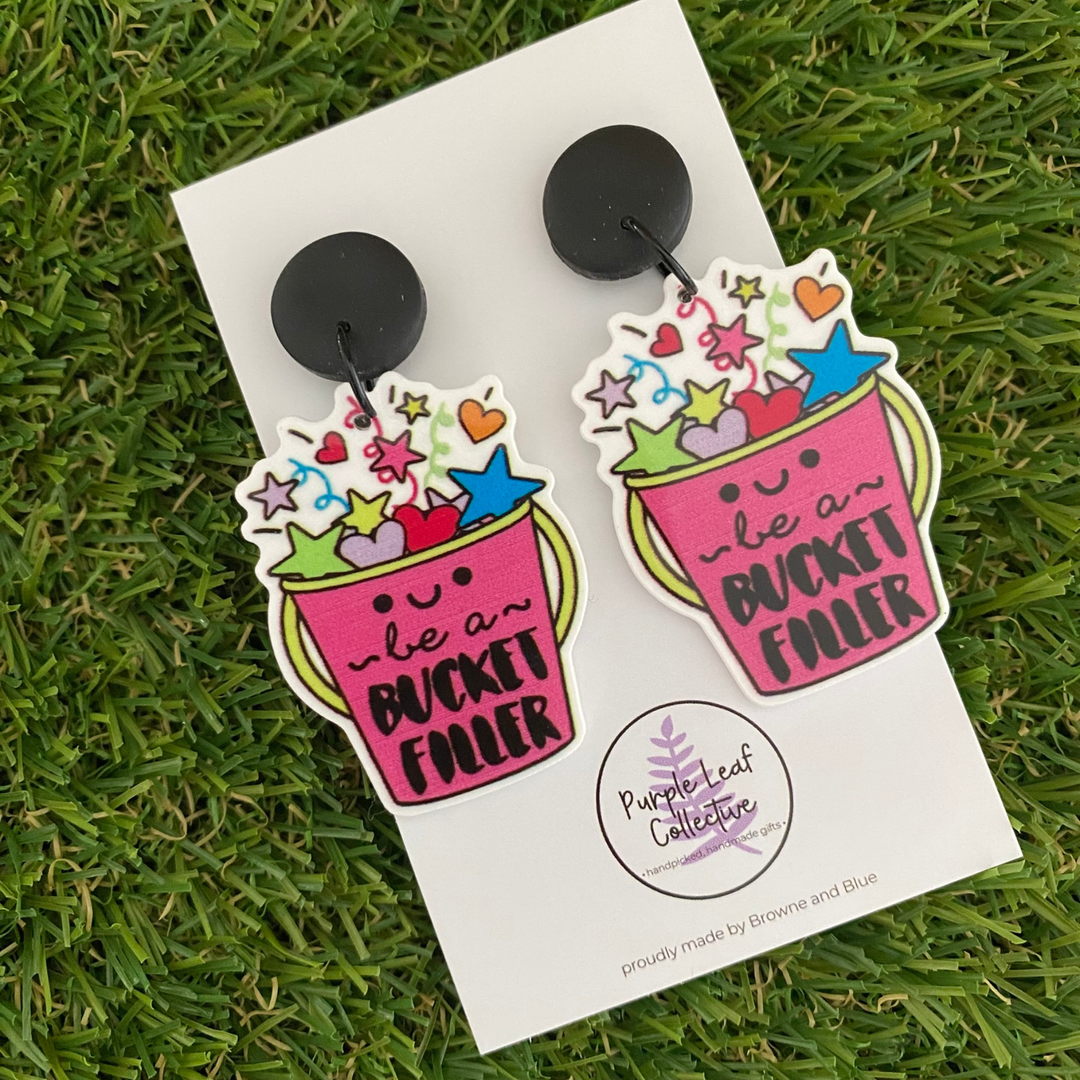 A pair of dangle earrings featuring a vibrant raspberry-coloured bucket adorned with colourful stars, hearts, and the uplifting phrase, set on black clay tops.