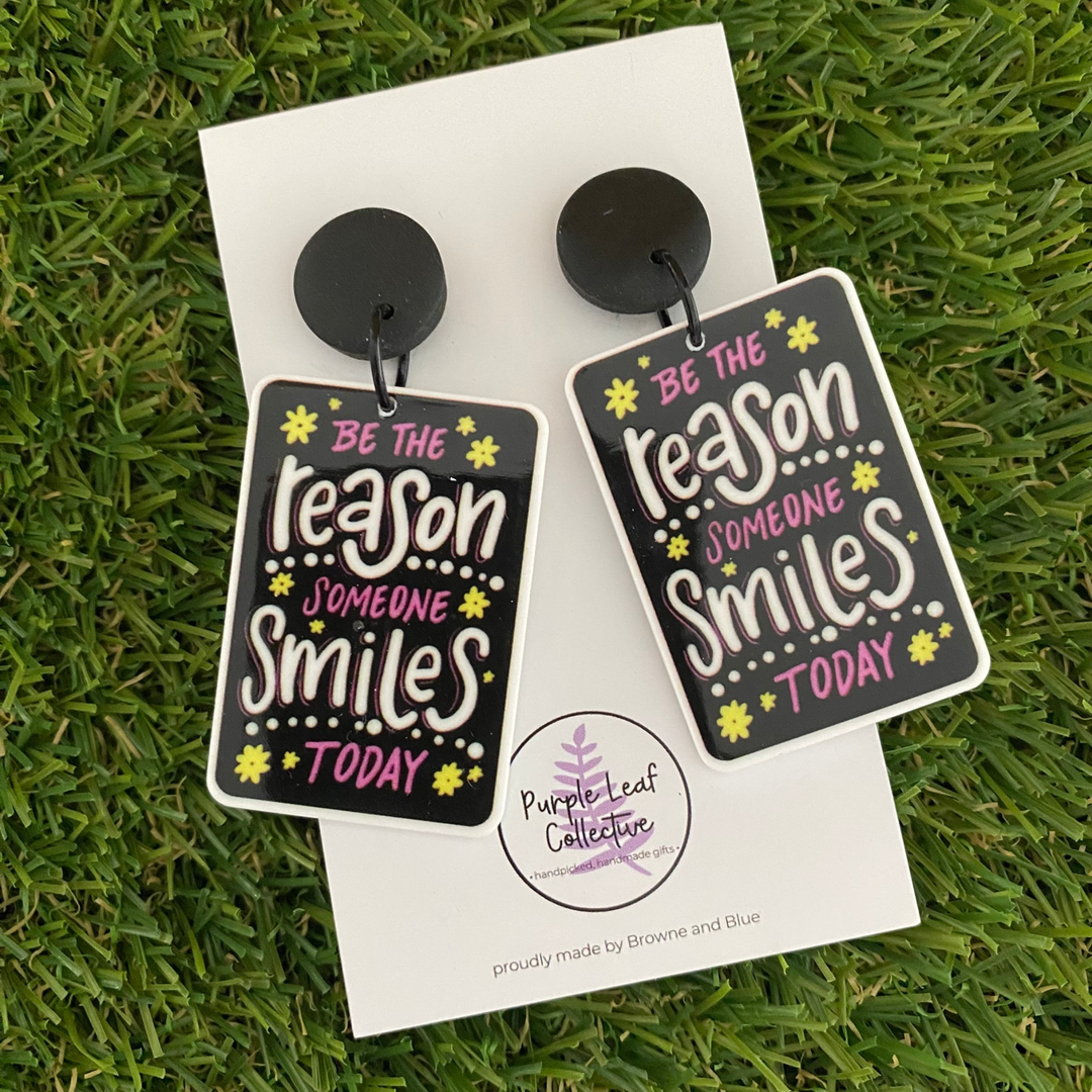 A pair of dangle earrings featuring two black rectangles with the inspiring wording set on sleek black clay tops.