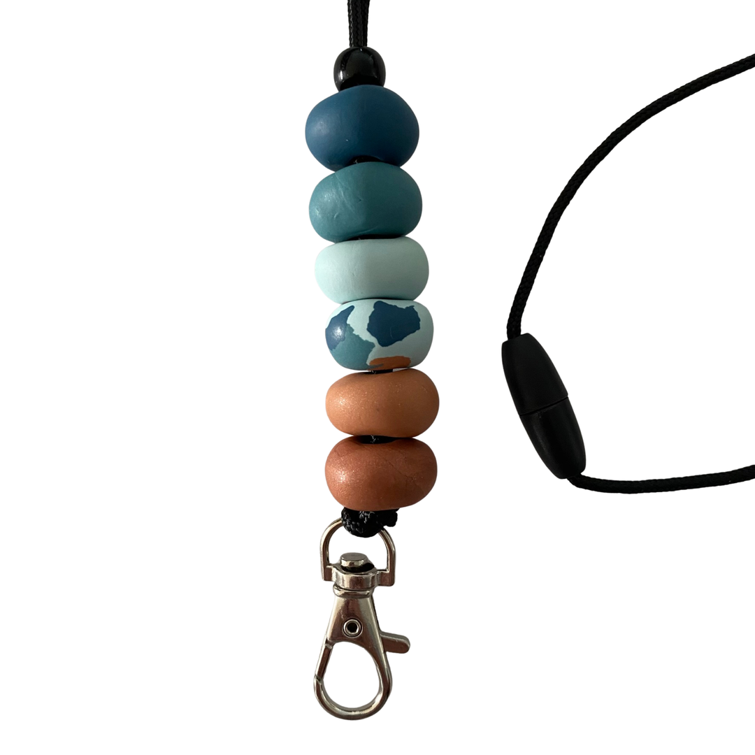Handmade polymer clay lanyard with six hand-rolled beads in sandy browns, ocean teals and pattern effects. Threaded on a nylon cord with a breakaway safety clasp and a silver-plated clip.