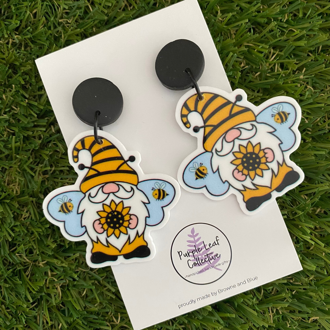 Pair of dangle earrings featuring gnomes wearing bee hats, holding a sunflower with bees in the background, set on sleek black clay tops.