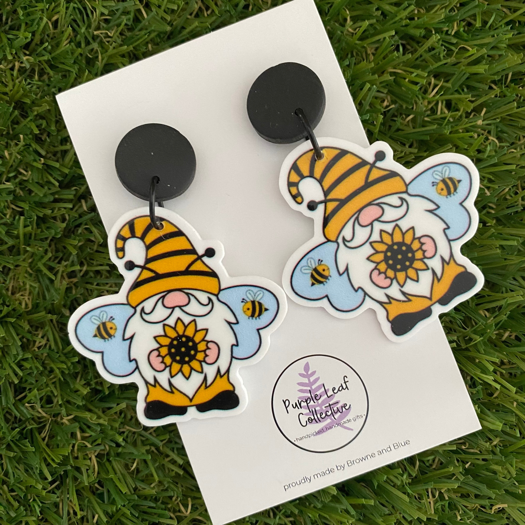 Pair of dangle earrings featuring gnomes wearing bee hats, holding a sunflower with bees in the background, set on sleek black clay tops.