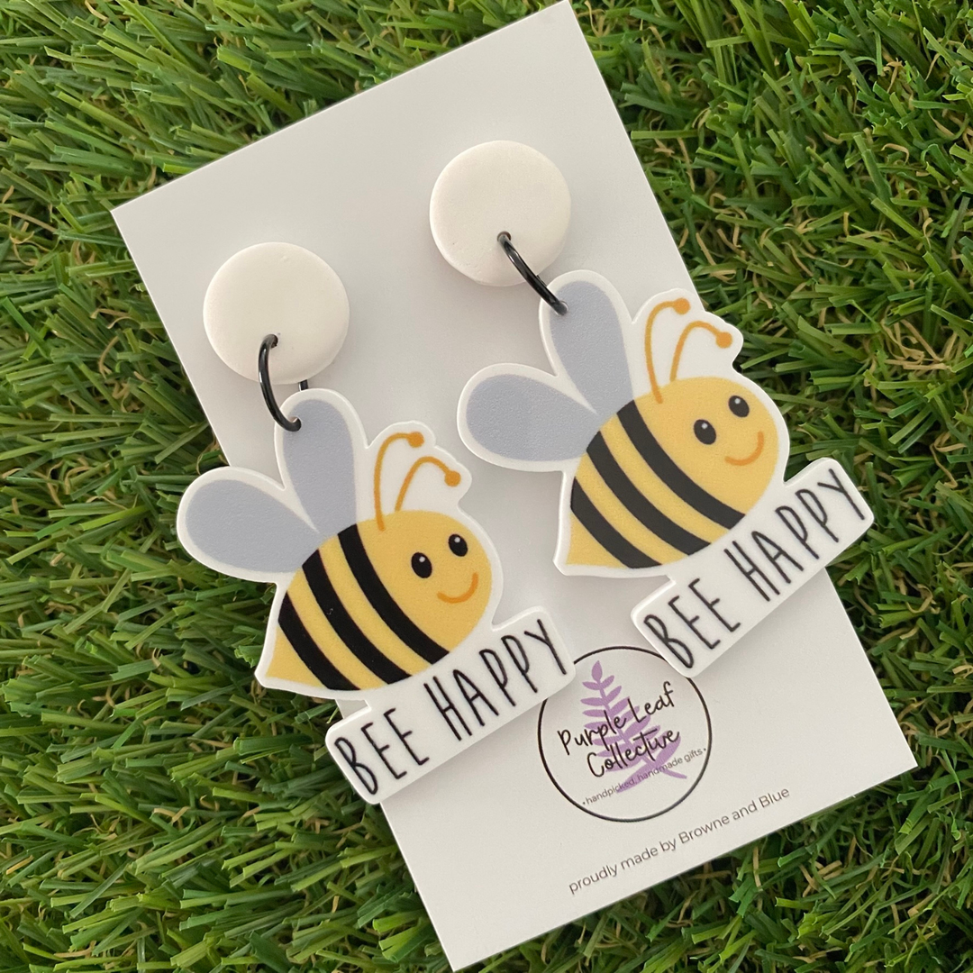 A pair of bee dangle earrings with the words BEE HAPPY on white stud tops.