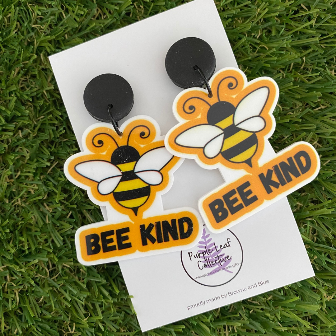 A pair of dangle earrings featuring two bees with the words Bee Kind underneath, set on black clay tops.