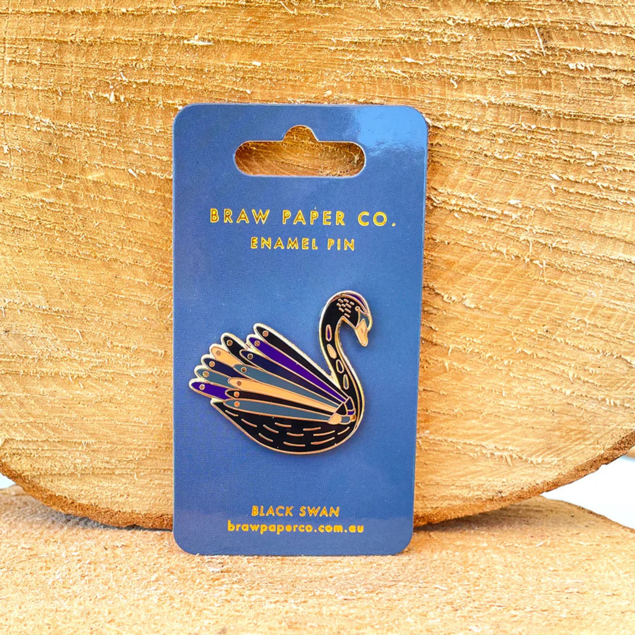 Enamel pin of a black swan on a blue card with 'Braw Paper Co.' branding, against a wooden background.