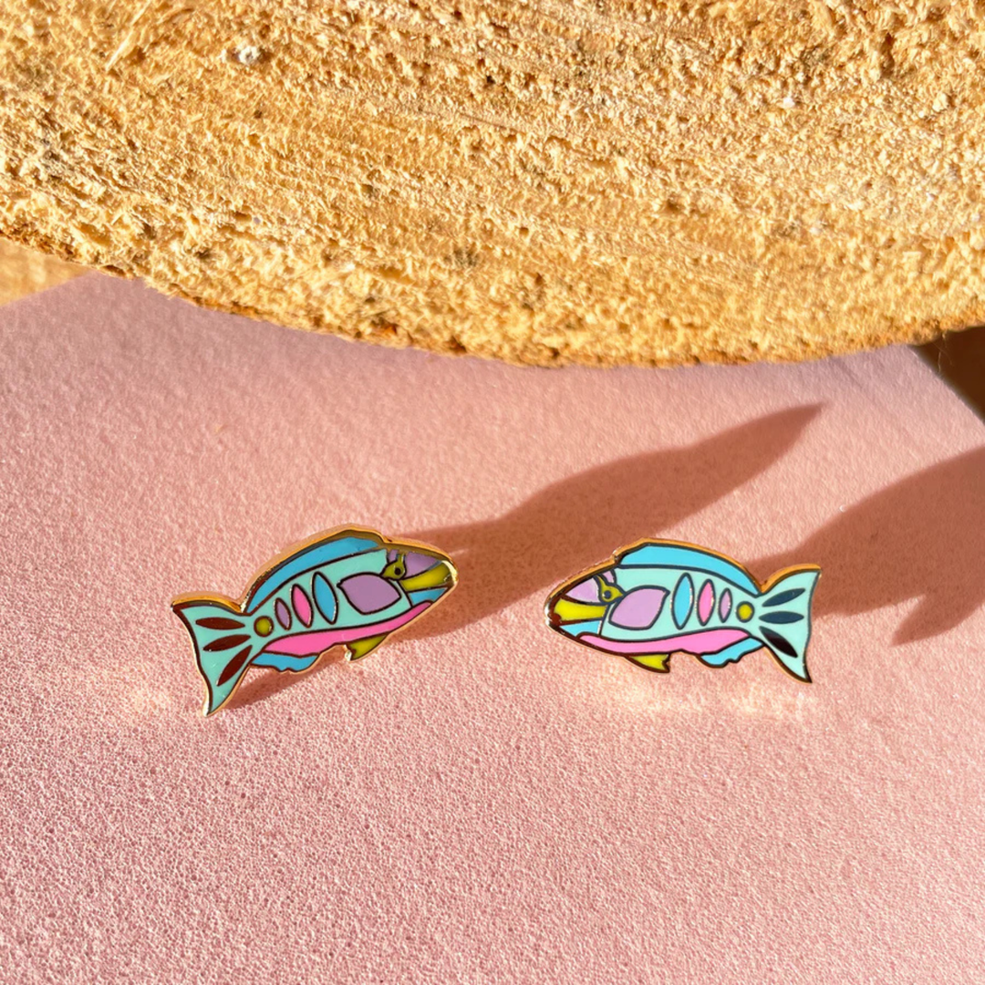 Colourful fish-shaped earrings on a pink surface with a gold rock in the background.