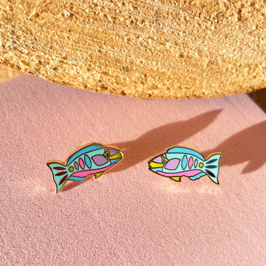 Colourful fish-shaped earrings on a pink surface with a gold rock in the background.