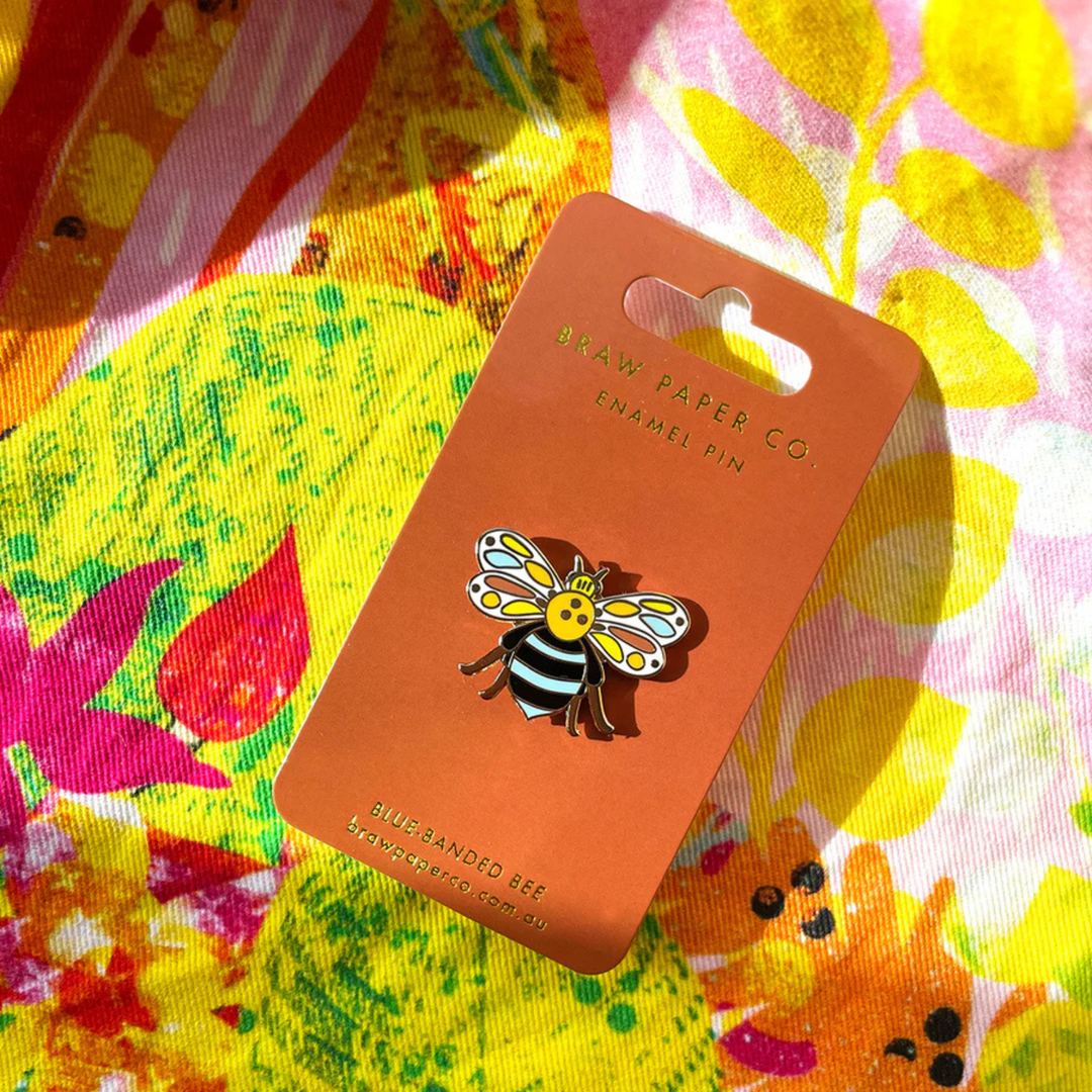 Enamel pin of a bee on an orange card with a colourful floral background.