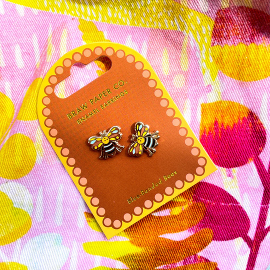 A pair of Blue-banded Bee enamel earrings on a Braw Paper Co. card with a floral background. 