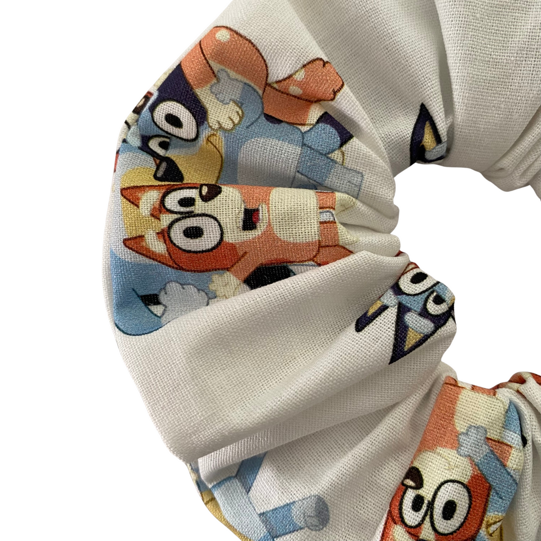 Close up view of a scrunchie featuring playful images of Bluey, Bingo, Bandit and Chilli on a white fabric.
