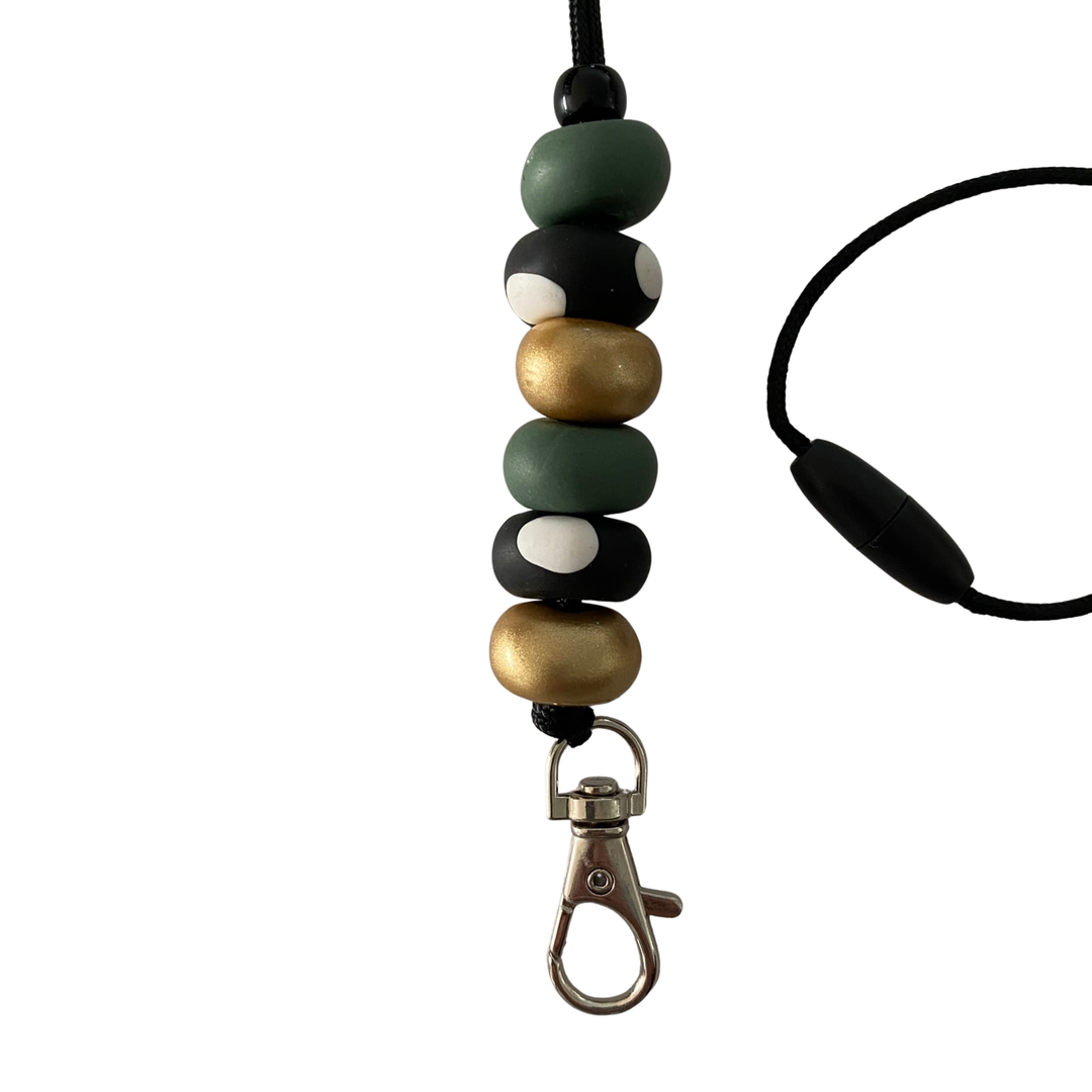 Close-up of six unique, handmade polymer clay beads in forest green, gold and spotted black beads on a lanyard cord with a breakaway safety clasp and a silver-plated clip.