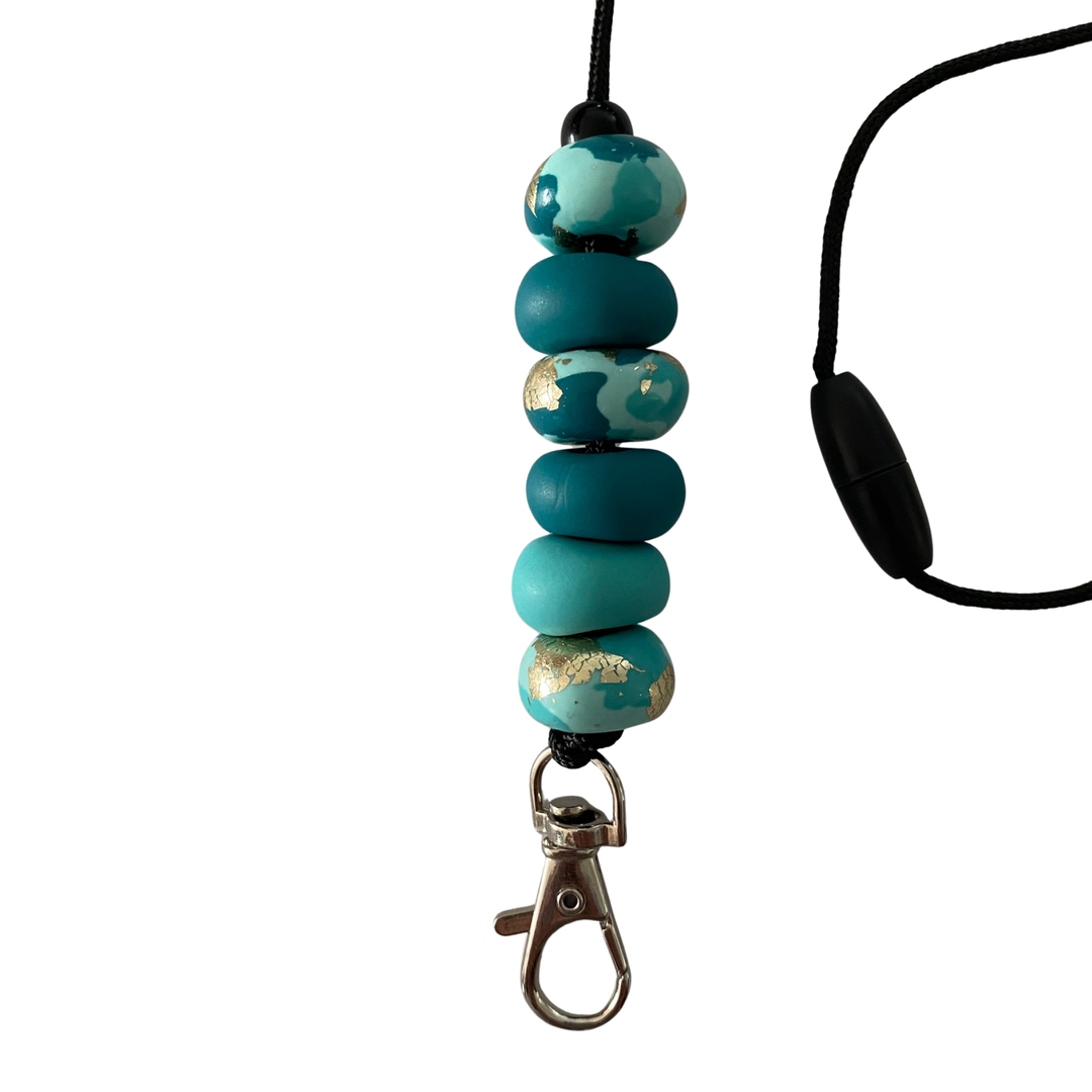 Close-up of six unique, handmade polymer clay beads in teals and silver foil on a lanyard cord with a breakaway safety clasp and a silver-plated clip.