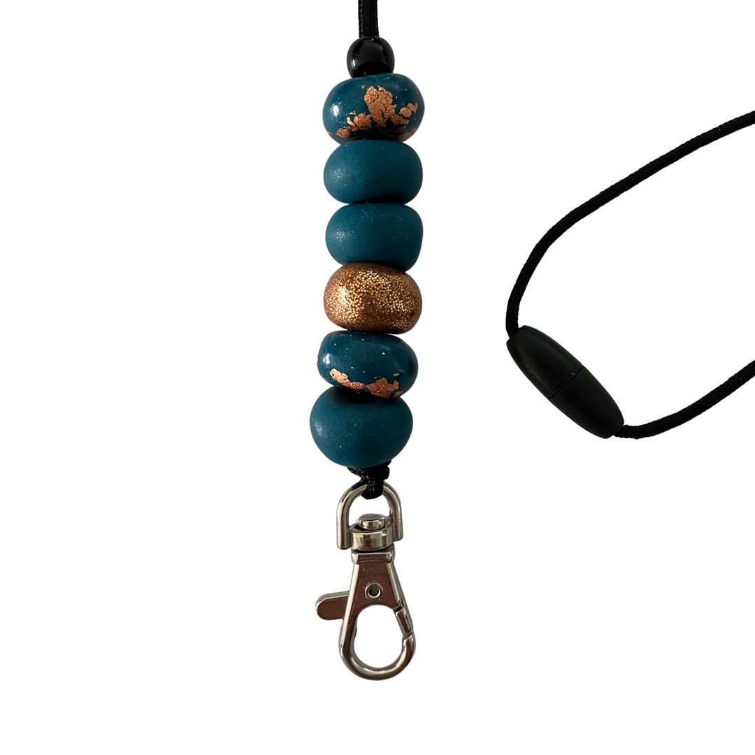 Handmade polymer clay lanyard with six hand-rolled beads in dark teal and gold colours. Threaded on a nylon cord with a breakaway safety clasp and a silver-plated clip.