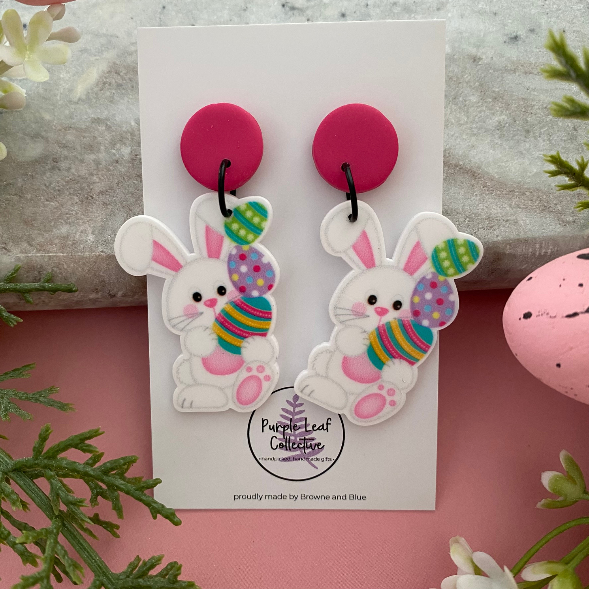 A pair of handmade dangle earrings featuring a white bunny balancing three colourful stacked Easter eggs with a pink stud topper.