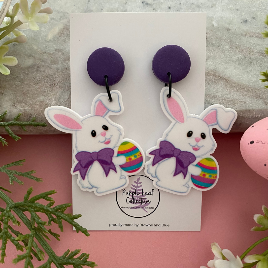 A pair of handmade dangle earrings featuring a white bunny wearing a purple bow and holding a colourful Easter egg with a purple stud topper.