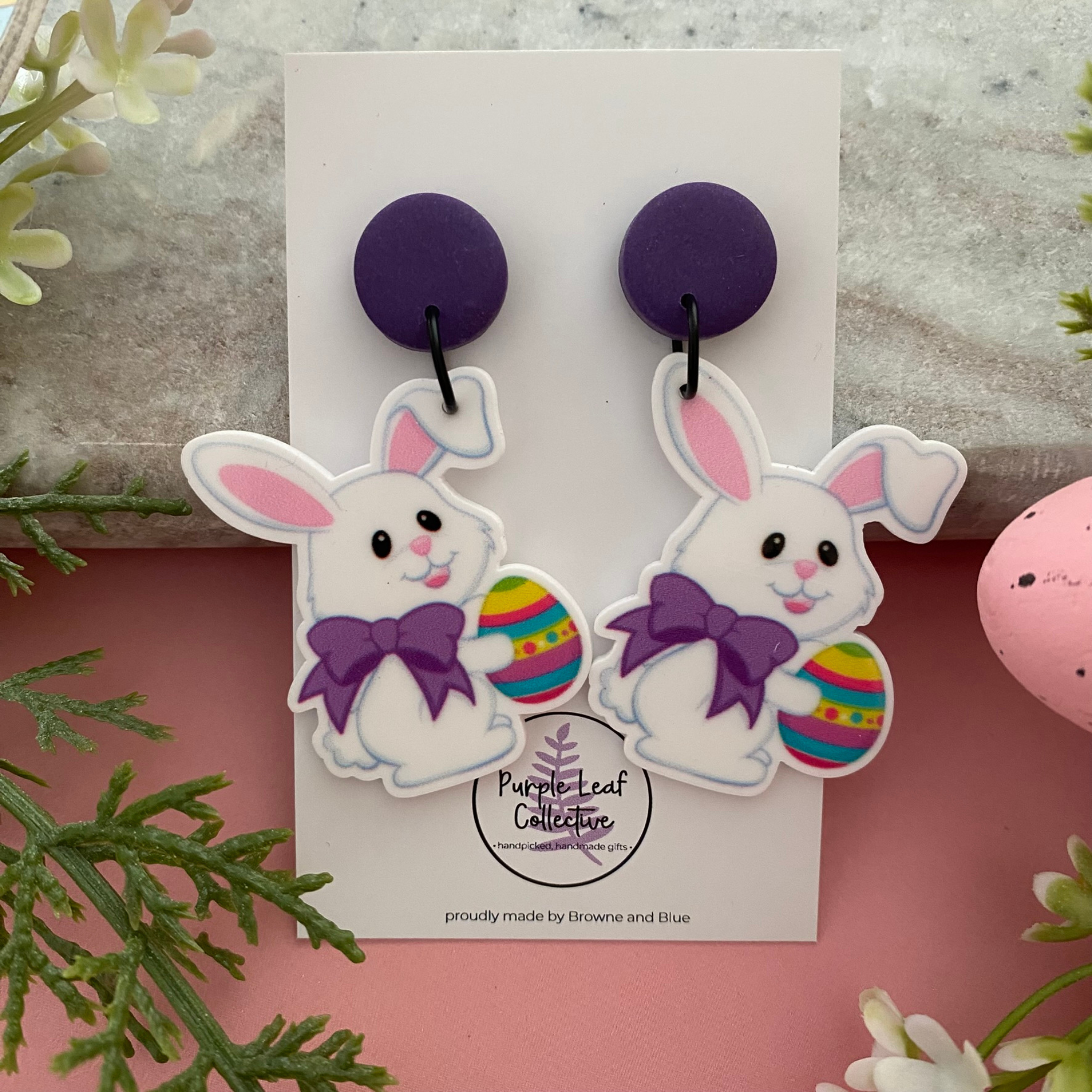 A pair of handmade dangle earrings featuring a white bunny wearing a purple bow and holding a colourful Easter egg with a purple stud topper.