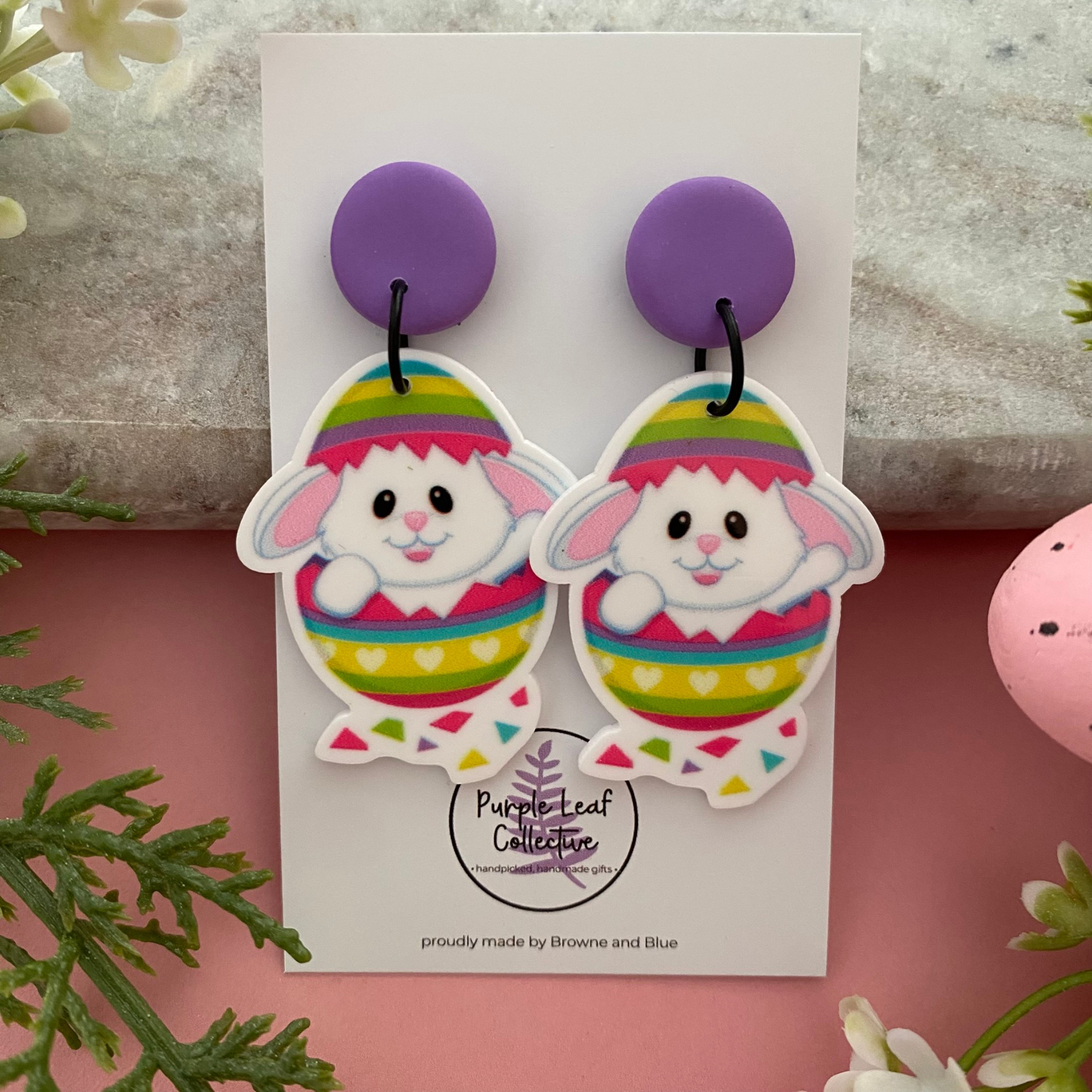 A pair of dangle earrings featuring a white bunny peeking out of a colourful striped Easter egg with purple stud top.
