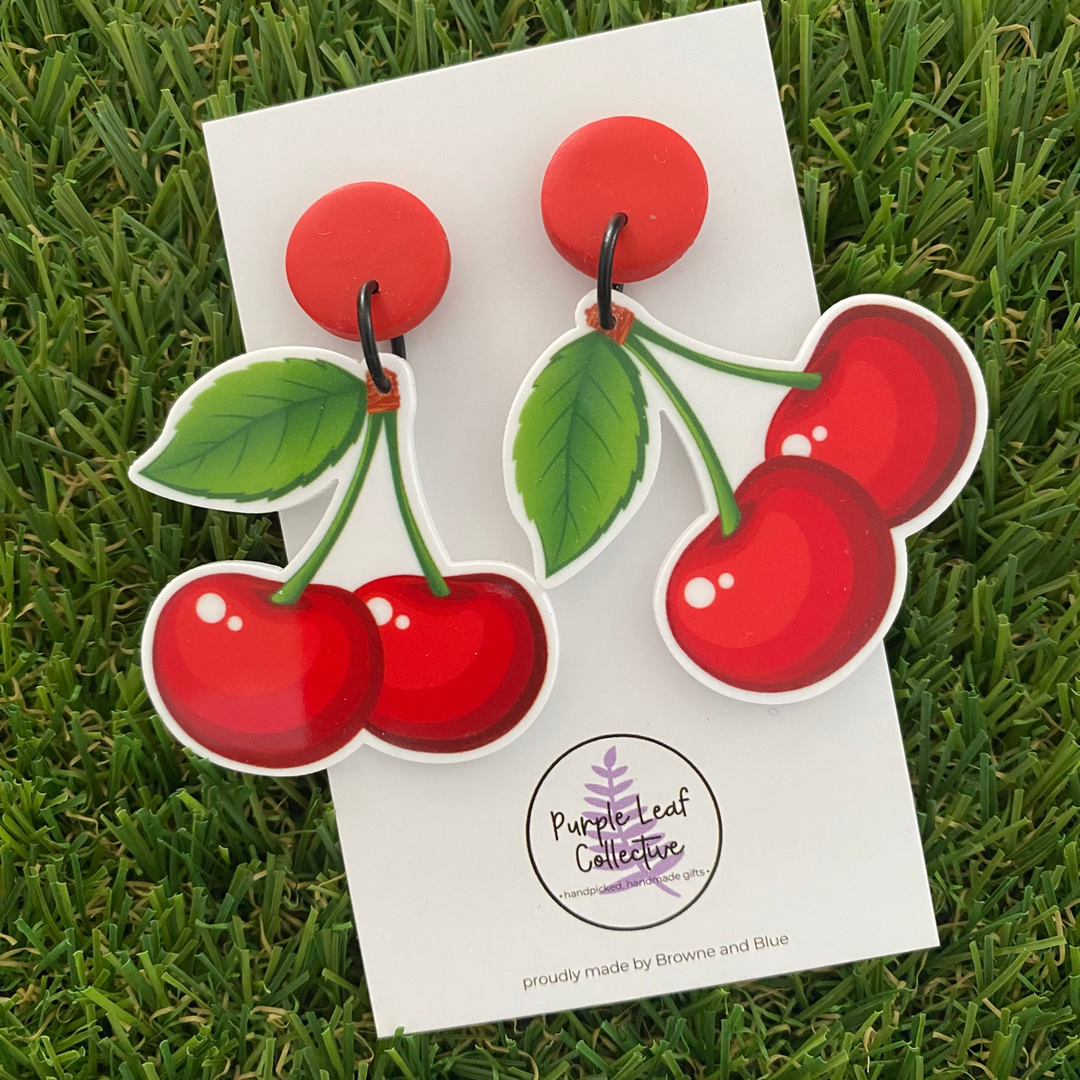 A pair of cherry-shaped earrings featuring two glossy red cherries with green stems and leaves, attached to red stud tops.