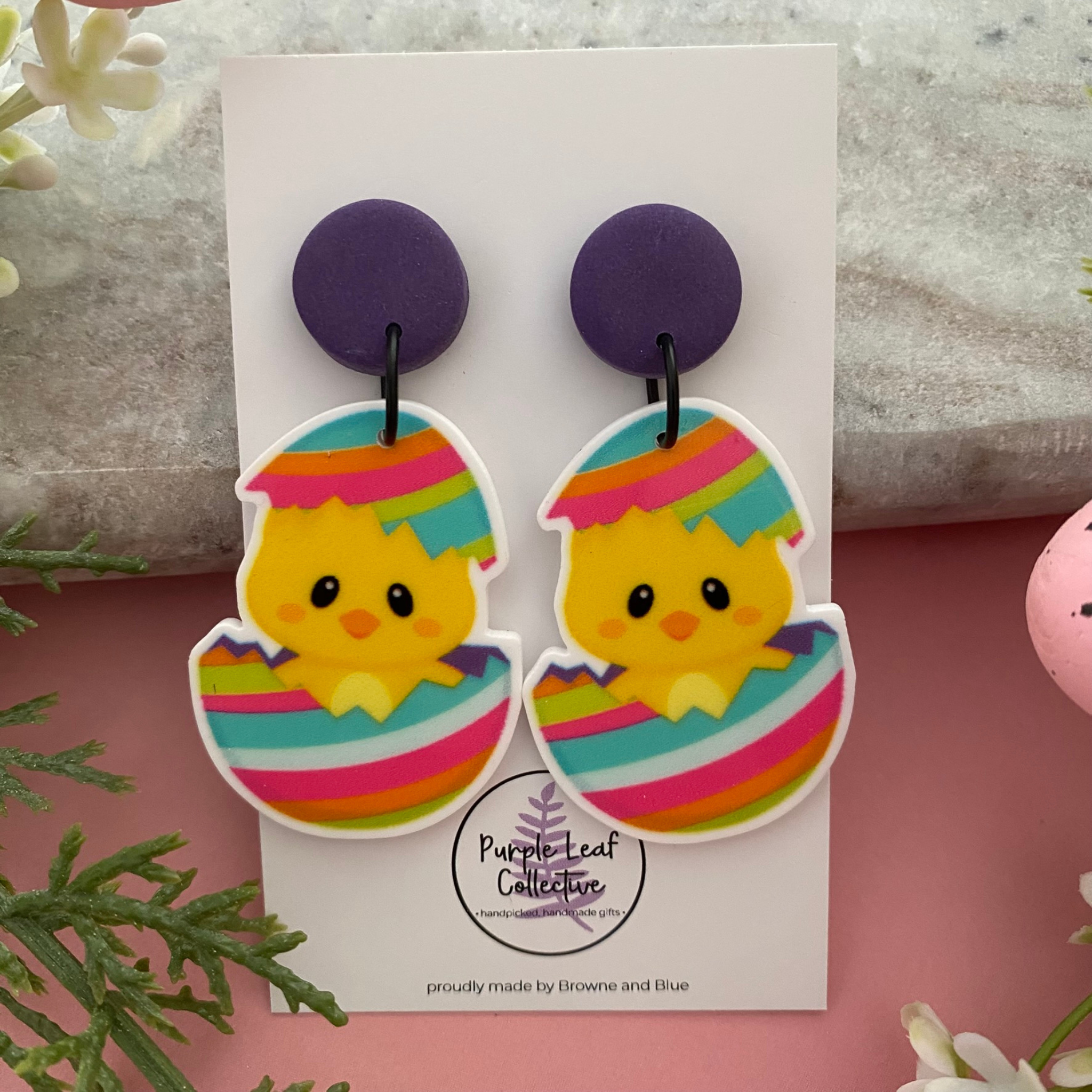 A pair of handmade dangle earrings featuring a sweet yellow chick peeking out of a colourful striped Easter egg, designed for lightweight comfort.