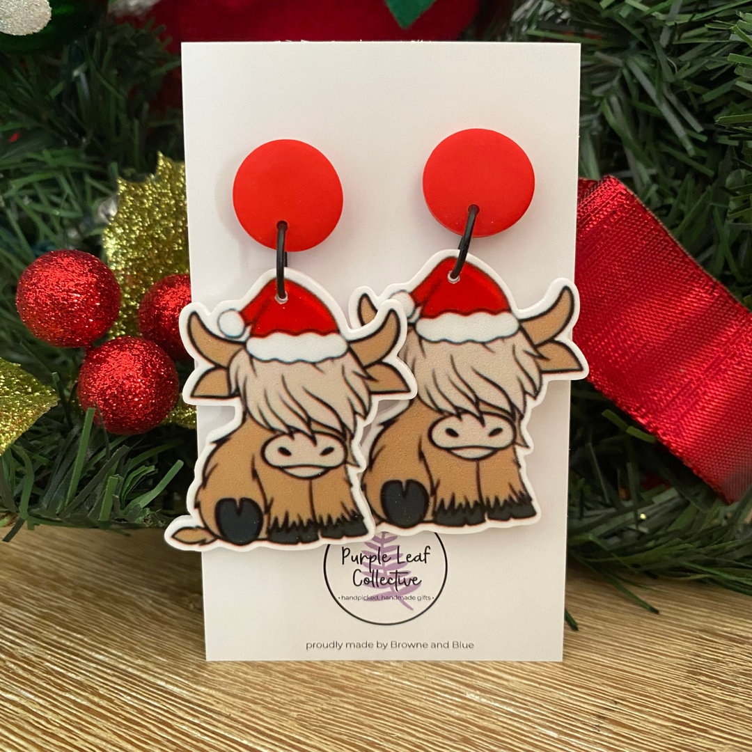 Pair of earrings with highland cows wearing Santa hats on a card with Christmas decorations in the background.