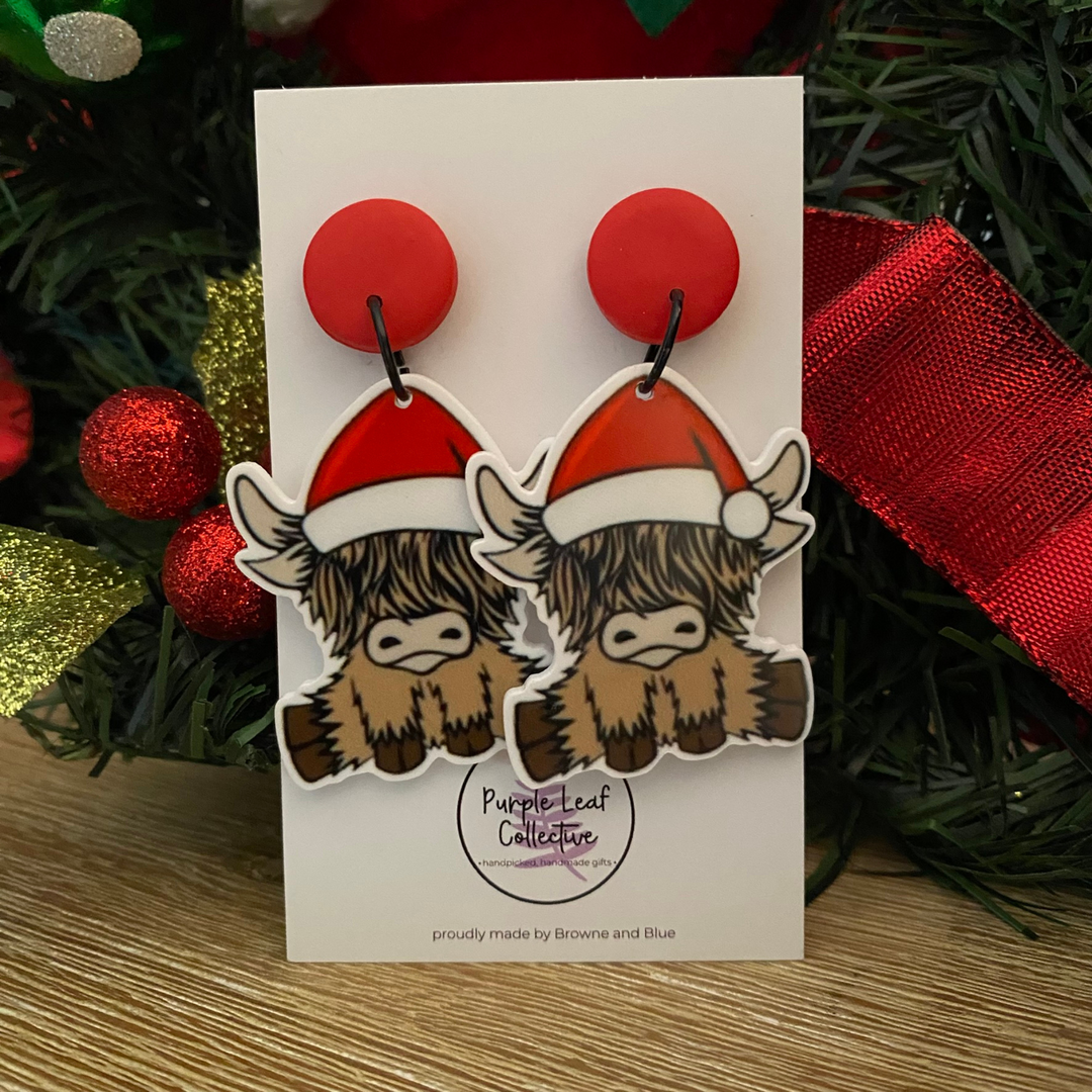 Pair of earrings with highland cows wearing Santa hats on a card with Christmas decorations in the background.