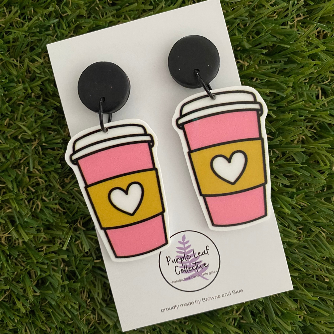 A pair of dangle earrings featuring a pair of pink coffee cups with white lids and love-heart sleeves set on sleek black clay tops.