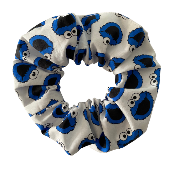 A handmade white cotton hair scrunchie featuring a repeated print of the blue Cookie Monster face. The scrunchie is bunched up, showing its texture and vibrant blue and white colour palette.