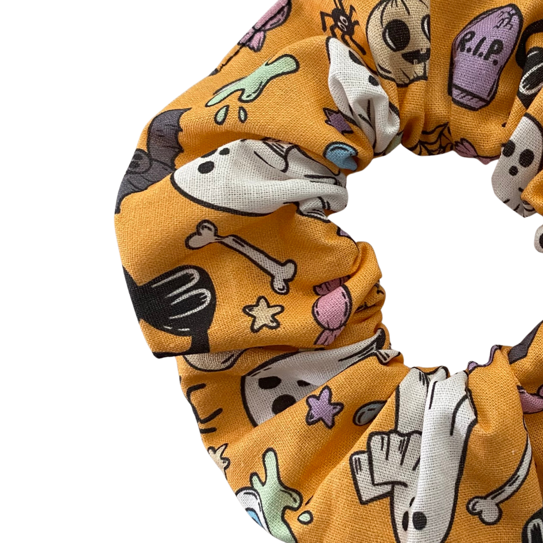 Close up view of a Halloween scrunchie featuring adorable collage of ghosts, pumpkins, coffins, cauldrons, bats and black cats on a light orange fabric.