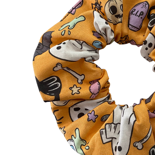 Close up view of a Halloween scrunchie featuring adorable collage of ghosts, pumpkins, coffins, cauldrons, bats and black cats on a light orange fabric.