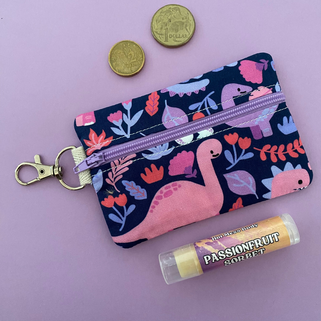 A small clip coin purse perfect for coins and small essentials featuring playful illustrations of iconic dinosaurs like T-Rex, Triceratops, and Brachiosaurus set against a vibrant purple background.