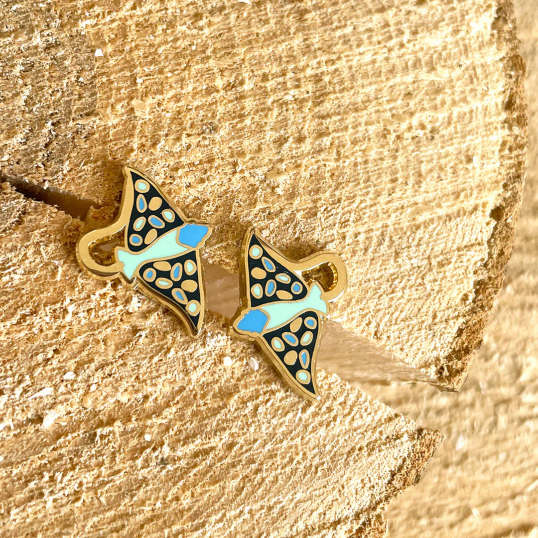 A pair of eagle ray enamel stud earrings with blue accents on a textured gold surface.