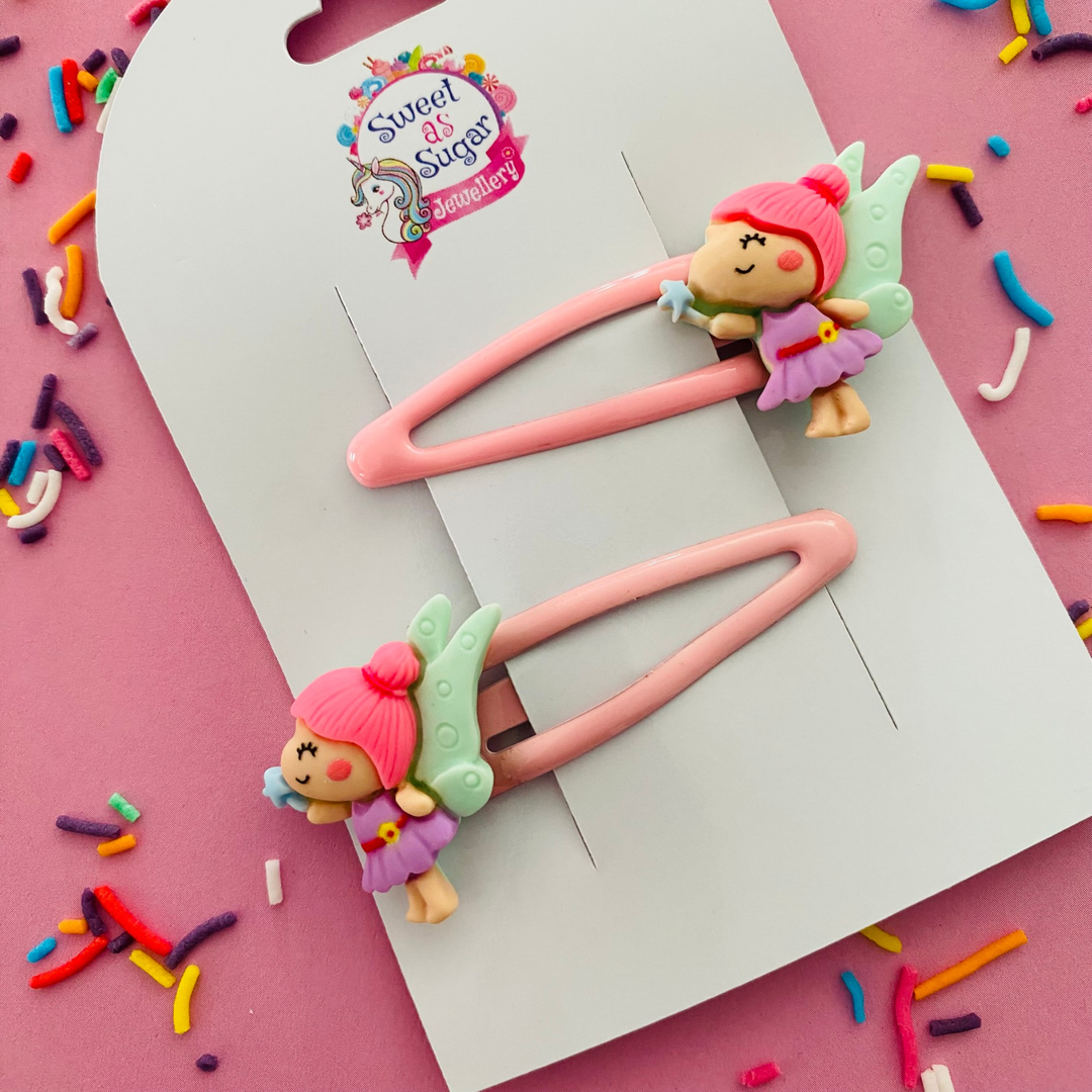 A pair of pink snap hairclips with fairy charms featuring green wings and blue wand.