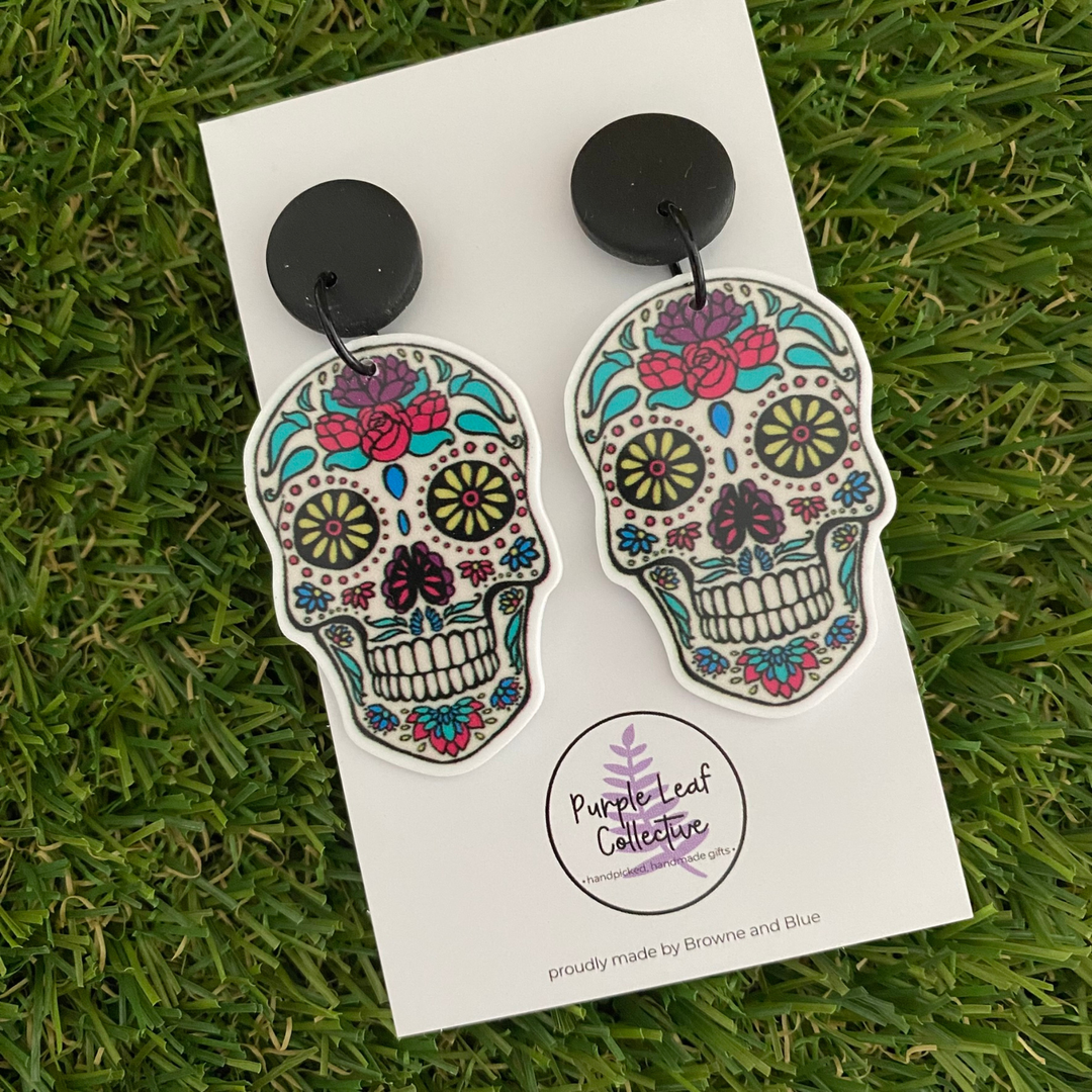 Colourful skull-shaped earrings set on black clay studs presented on a white backing card.
