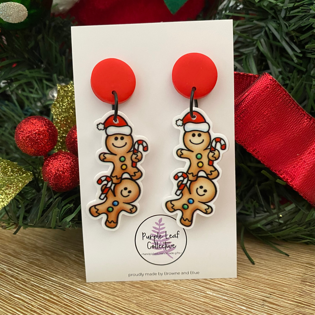 Pair of gingerbread man earrings on a card with Christmas decorations in the background.