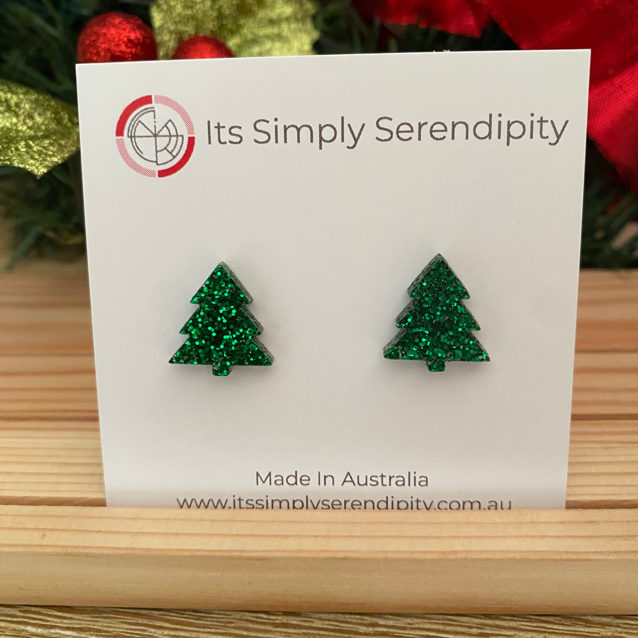 Green glittery tree-shaped earrings on a 'Its Simply Serendipity' card with Christmas decorations in the background.