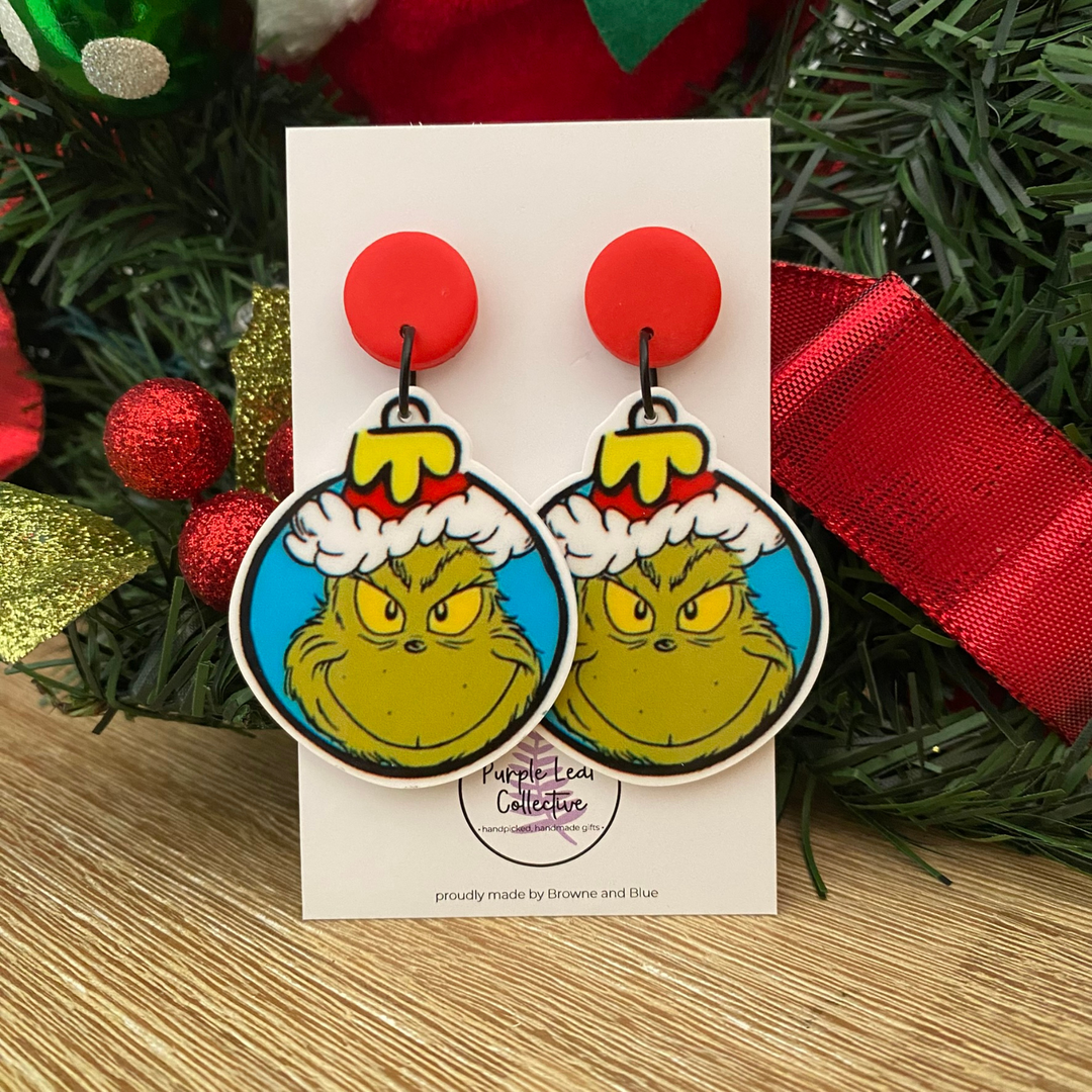 Pair of Grinch earrings on a card with Christmas decorations in the background.