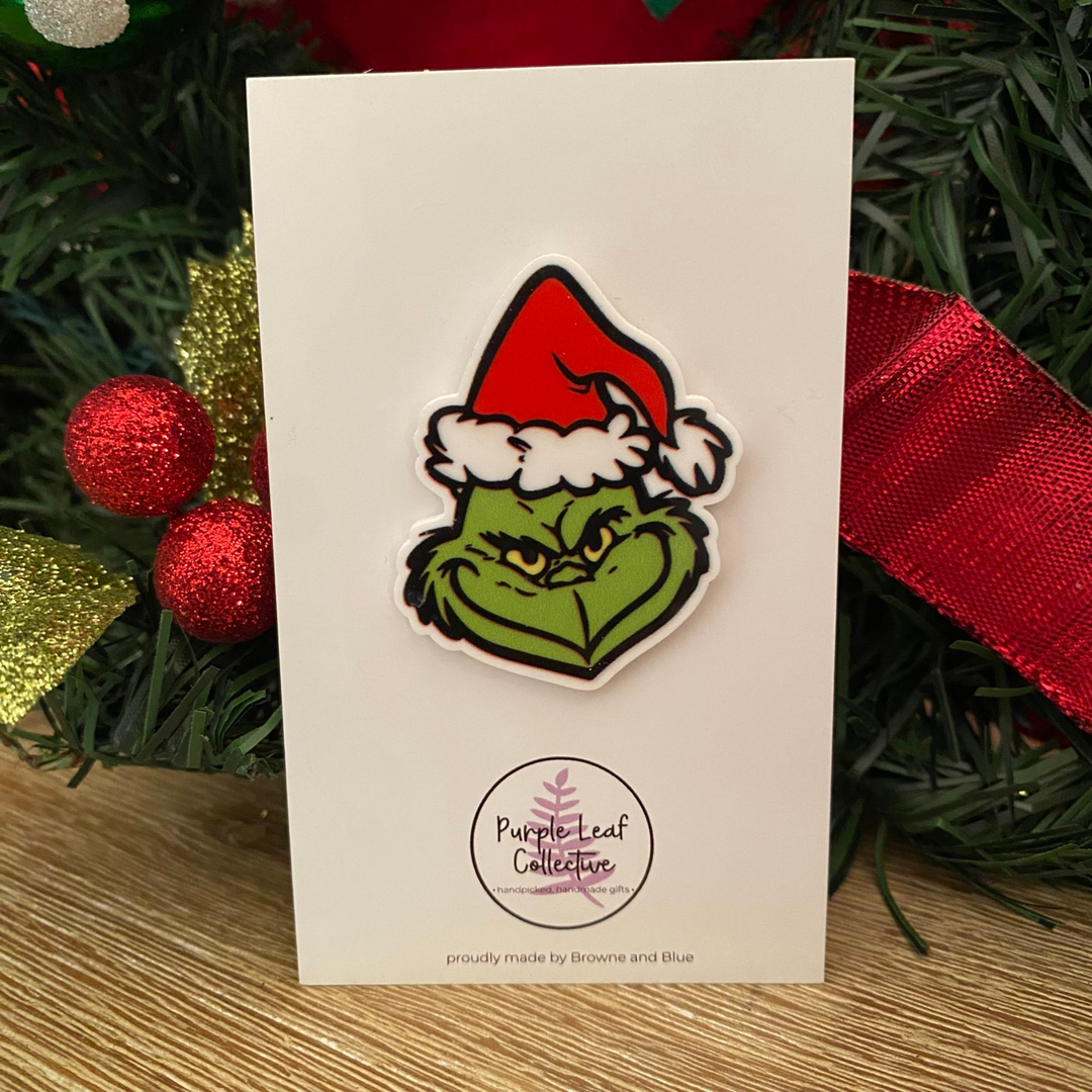 Grinch Christmas-themed brooch on a card with decorative background.