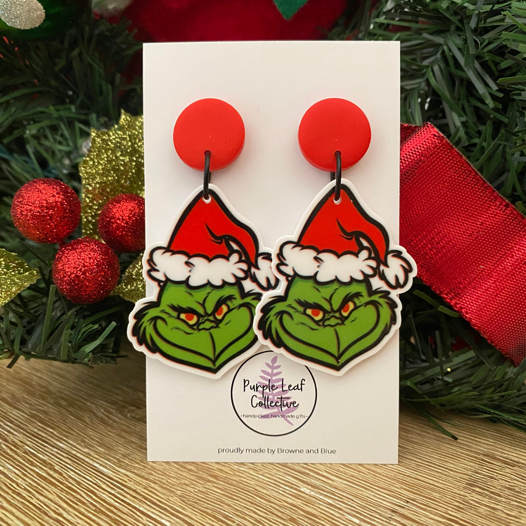 Grinch earrings on a card with Christmas decorations in the background.