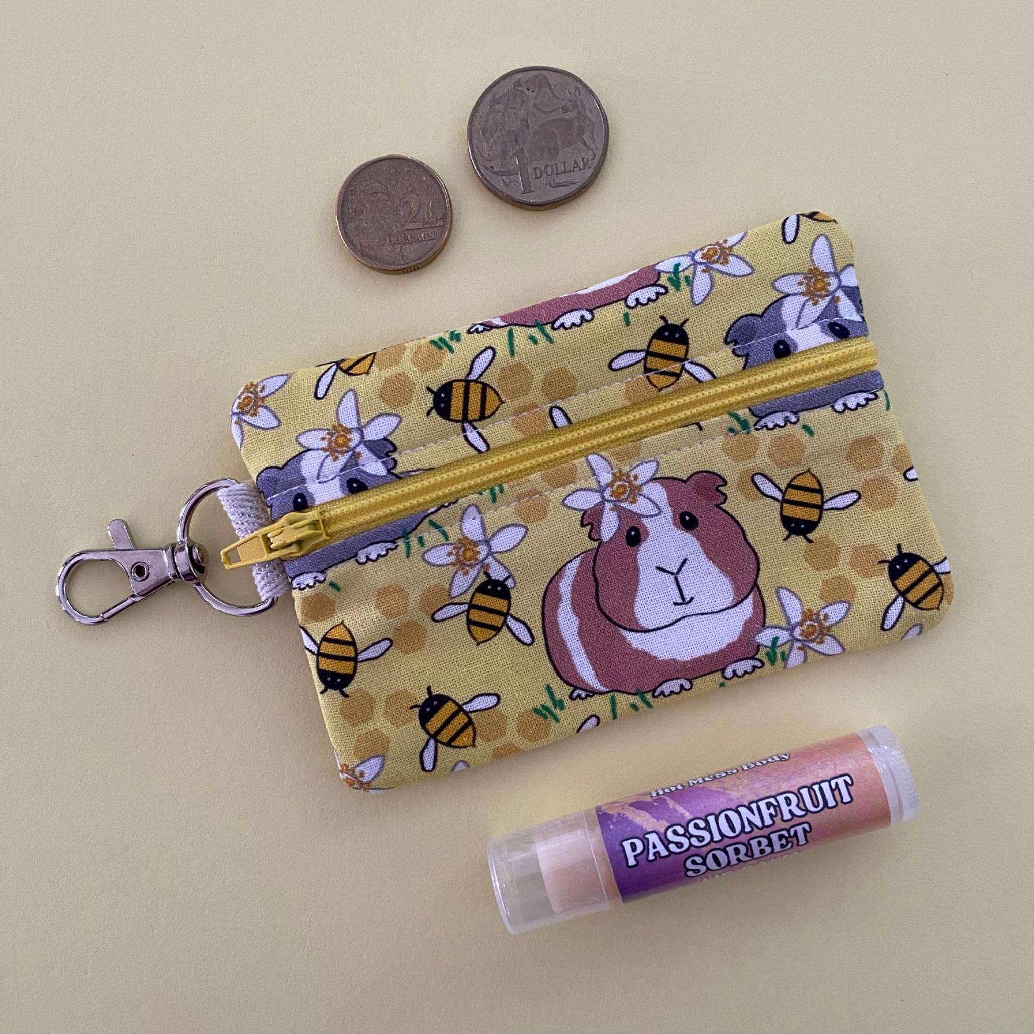 Small pouch with guinea pig and bee pattern, lip balm, and coins on a beige background.