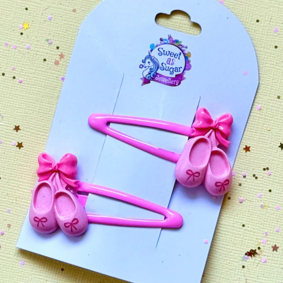 A pair of pink ballet shoe hair clips. The charms feature tiny ballet slippers with a pink bow, mounted on a matching pastel pink metal snap clip.