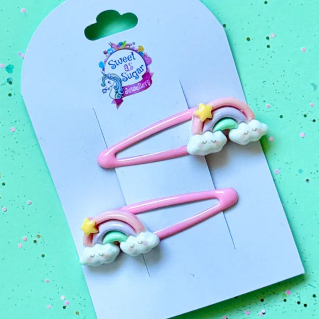 A hair clip featuring a pastel-coloured rainbow arch resting on two fluffy white clouds with a small yellow star, attached to a pink metal snap clip.