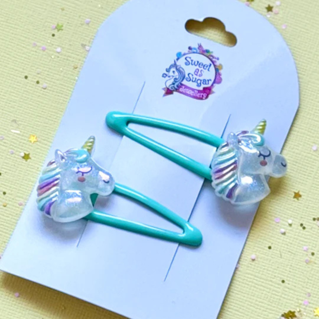 A unicorn-themed hair clip featuring a unicorn charm with a golden horn, attached to a bright aqua-blue snap clip.