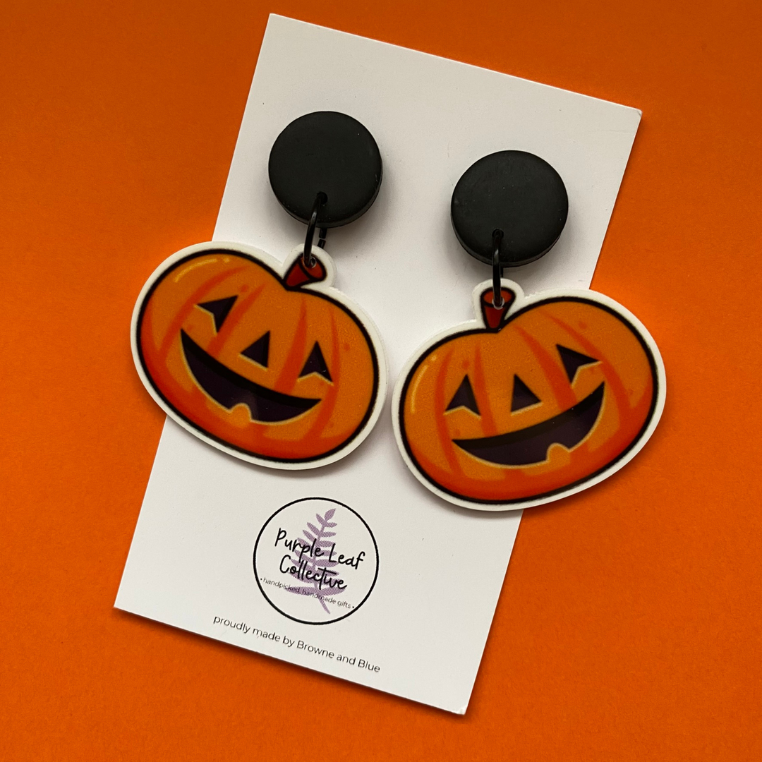 Halloween-themed pumpkin earrings on a card with 'Purple Leaf Collective' branding against an orange background.